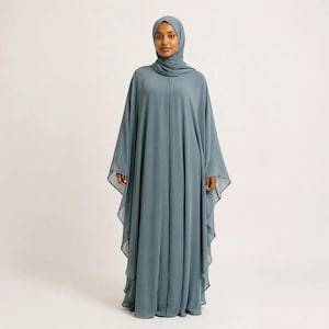 Puede incluir: Una abaya de color azul pizarra que llega hasta el suelo, con una silueta holgada y fluida, mangas anchas y drapeadas, y un pañuelo a juego. La prenda está confeccionada con una tela ligera de color sólido que cae en pliegues verticales.