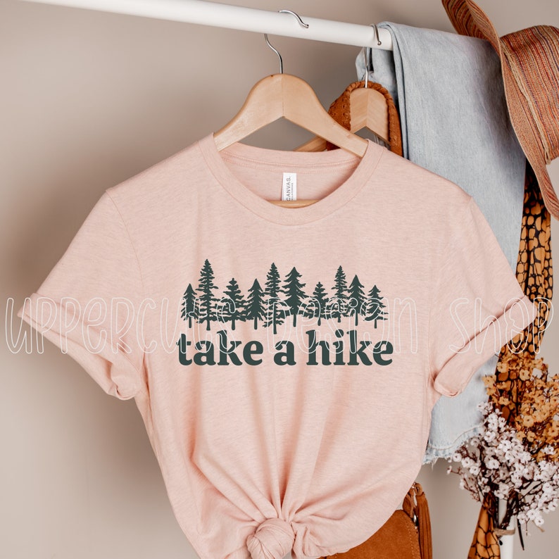 Op de afbeelding: Een roze T-shirt met een afbeelding van een rij dennenbomen en de tekst "take a hike" in zwart.