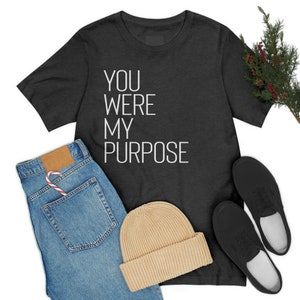 Peut inclure: Un t-shirt gris foncé avec un texte blanc qui dit "You were my purpose". Le t-shirt est posé à plat sur un fond blanc avec une paire de baskets noires, un bonnet et un jean bleu.