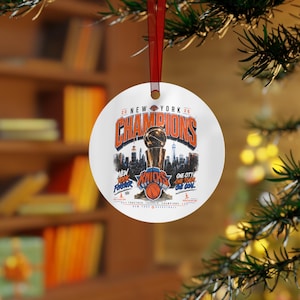 May include: A round white ceramic ornament hanging from a red ribbon. The design features the text New York Champions, a basketball trophy, a New York City skyline silhouette, and the New York Knicks logo with a basketball and the text New York Forever.