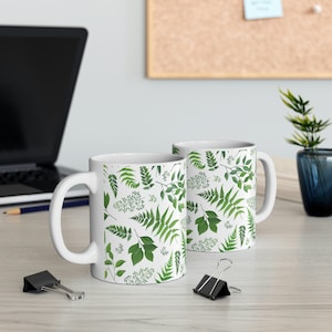 May include: Two white ceramic mugs with a green leafy pattern. The mugs have a white background with a repeating pattern of green leaves and ferns.