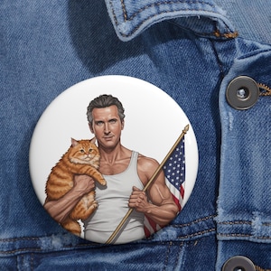 Può includere: Un bottone bianco con un uomo muscoloso in canottiera che tiene un gatto arancione e una bandiera americana. L'uomo ha i capelli corti e grigi e un'espressione seria. Il gatto è arancione e soffice.