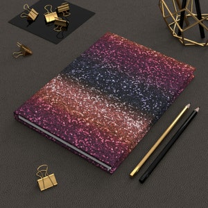 May include: A pink, orange and blue glitter notebook with a black spine. The notebook is lying on a dark brown surface with two pencils and gold paper clips.