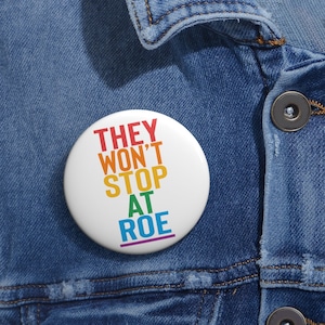 Peut inclure: Un bouton blanc avec le texte "THEY WON'T STOP AT ROE" en couleurs arc-en-ciel. Le bouton est épinglé sur une veste en jean bleue.
