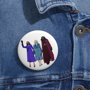 May include: A white button with a colorful illustration of three women wearing face masks and coats. The women are standing side-by-side, with the woman on the left waving. The woman in the middle is wearing a blue coat, the woman on the right is wearing a red coat, and the woman on the left is wearing a purple coat.