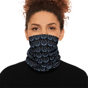 May include: Black neck gaiter with a repeating blue and black graphic pattern. The pattern features a stylized symbol. The neck gaiter is worn around the neck and covers the lower face.