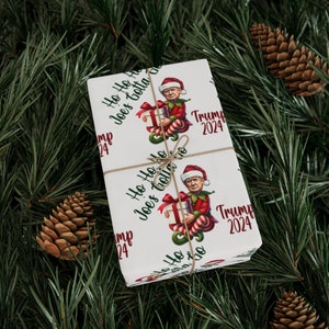 Può includere: Carta da regalo bianca con un motivo ripetuto di un elfo dei cartoni animati che tiene una scatola regalo. L'elfo indossa un cappello rosso e un costume verde e rosso. Il testo "Ho Ho Ho Joe's Gotta Go" e "Trump 2024" è stampato sulla carta.