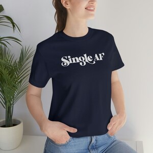 Puede incluir: Una camiseta azul marino con texto blanco que dice "Single AF" en una fuente retro.