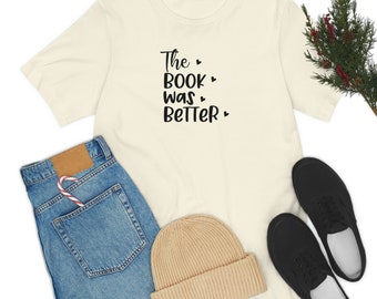 Pode incluir: Camiseta creme com o texto preto "The Book Was Better" e corações. Combinada com jeans azuis, um gorro bege, sapatos pretos e uma bengala de doces no bolso.