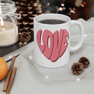 May include: White ceramic mug with a pink heart-shaped design that says "LOVE" in a retro font. The mug is filled with coffee and sitting on a white surface with string lights.