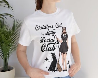 Op de afbeelding: Wit T-shirt met de tekst "Childless Cat Lady Social Club" in zwart schrift. De afbeelding toont een cartoonvrouw in een zwarte jurk met een bezem, een zwarte kat en twee sterren. Het shirt is gemaakt van een zacht materiaal.