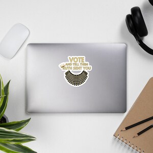 Op de afbeelding: Een witte laptop sticker met een gouden kroon, een zwarte kraag en de tekst "VOTE AND TELL THEM RUTH SENT YOU".