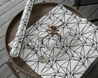 May include: White wrapping paper with a black geometric pattern. A brown ribbon is tied around a white gift box. Scissors and a roll of wrapping paper are also on the table.