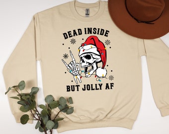 Könnte beinhalten: Beiges Sweatshirt mit einem Totenkopf-Motiv, der eine Weihnachtsmütze und Lichterketten trägt. Der Text lautet "DEAD INSIDE BUT JOLLY AF". Ein brauner Hut liegt oben rechts auf dem Sweatshirt.