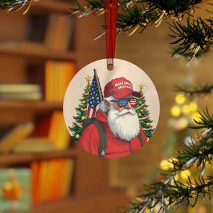 May include: A round Christmas bauble depicting a cartoon Santa Claus wearing a red "Make America Great Again" hat and sunglasses. He is standing in front of a Christmas tree with an American flag behind him.