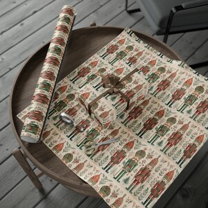 May include: A roll of wrapping paper with a repeating pattern of nutcracker soldiers, flowers, and greenery on a cream background. A smaller gift box is wrapped in the same paper and tied with a brown ribbon. The gift box is sitting on a wooden tray.