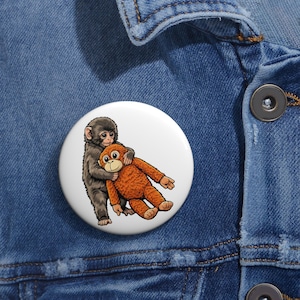 May include: A white button featuring a cartoon illustration of two monkeys hugging. One monkey is gray, and the other is orange. The button is set against a blue denim jacket.