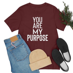 Peut inclure: Un t-shirt marron avec un texte blanc qui dit "YOU ARE MY PURPOSE". Le t-shirt est posé à plat sur un fond blanc avec une paire de chaussures noires, un jean bleu et un bonnet beige.