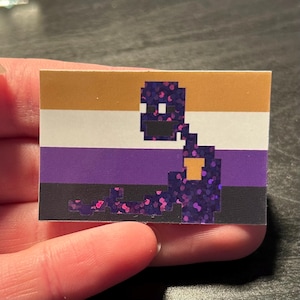 May include: A purple, black, and white pride flag with a pixelated purple character on top. The character has a black mouth and white eyes.
