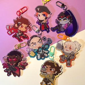 May include: A collection of seven acrylic keychains featuring stylized chibi characters with colorful plastic clasps. Each keychain displays a unique character design with distinct outfits, hair colors, and accessories, including a mug, a beret, and a beer stein.