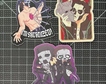 May include: Three stickers featuring illustrations of a band member. One sticker shows a shirtless figure with swords piercing a heart, with the text "I'M SATANIZED!". Another shows a figure holding a mask. The third shows two figures with skeleton-like features.