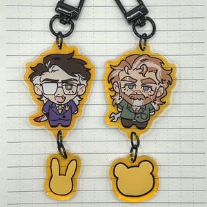 May include: Two cartoon character keychains with black metal clasps. The first keychain features a man with glasses wearing a purple suit and holding a bloody knife. The second keychain features a man with a beard wearing a green shirt and holding a wrench. Both keychains have a yellow charm attached to the bottom.