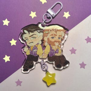 Può includere: Un portachiavi in acrilico trasparente con due personaggi dei cartoni animati con accenti glitter. I personaggi indossano gilet viola con accenti gialli e sono seduti uno accanto all'altro. Un ciondolo a forma di stella gialla pende dal portachiavi.