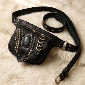 Può includere: Marsupio in pelle nera con borchie e dettagli color oro. La borsa presenta una pietra ovale e una patta decorativa. La cintura regolabile ha una chiusura con fibbia e un passante più piccolo. La borsa è su una superficie di pelliccia bianca.