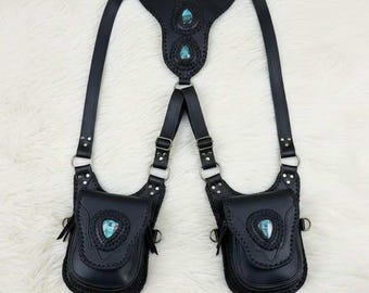 May include: A black leather harness featuring two symmetrical pouches attached to adjustable straps. The central back panel and each pouch flap are decorated with a teardrop-shaped turquoise stone. Silver-toned metal hardware connects the straps.