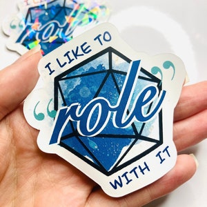 Puede incluir: Pegatina blanca con el texto "I LIKE TO role WITH IT" en azul. La palabra "role" está superpuesta a una forma geométrica azul y blanca. Una segunda pegatina es visible en el fondo.