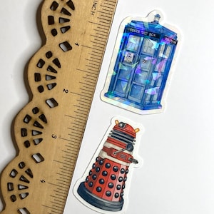 May include: Two stickers next to a wooden ruler showing a length of 5 inches. One sticker is a blue police box with holographic accents and the text Police Public Call Box. The other sticker is a red and black robotic figure with a dome top.