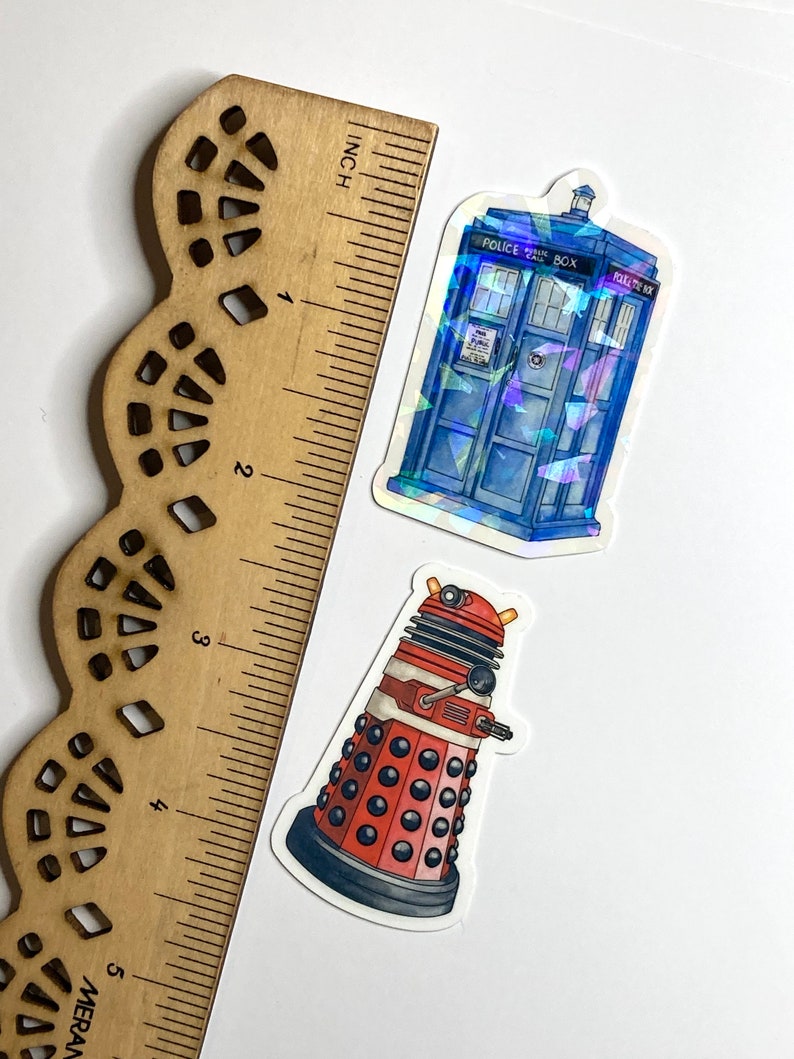 May include: Two stickers next to a wooden ruler showing a length of 5 inches. One sticker is a blue police box with holographic accents and the text Police Public Call Box. The other sticker is a red and black robotic figure with a dome top.