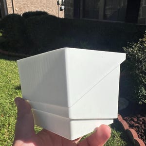 May include: A white plastic planter with a square shape and a slightly angled design. The planter is made of a smooth, white plastic and has a simple, modern look.