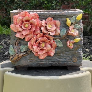 May include: A rectangular ceramic planter with a textured, wood-like surface. The planter is decorated with three dimensional pink flowers and green leaves.