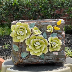 May include: A rectangular ceramic planter with a rustic brown glaze and a raised design of green and white flowers. The planter has a price tag that reads "$22.99".