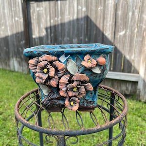 May include: A turquoise ceramic planter with raised floral details in shades of peach and brown. The square-shaped pot sits on a decorative, wrought-iron plant stand. The planter has a textured surface and is suitable for indoor or outdoor use.