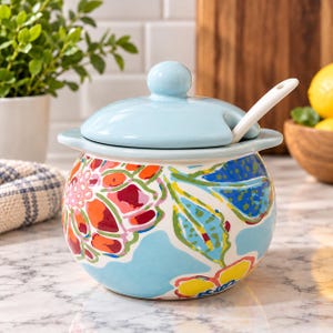 May include: A round ceramic sugar bowl with a light blue lid and a matching white spoon. The bowl features a vibrant floral pattern with red, orange, yellow, and blue colors on a white background. It has a small notch for the spoon handle.