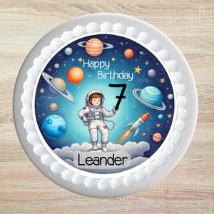 Könnte beinhalten: Ein runder Tortenaufleger mit Weltraummotiv, das einen Astronauten, Planeten, Sterne und eine Rakete zeigt. Der Text lautet Happy Birthday, die Zahl 7 und der Name Leander. Das Design ist von einem weißen Spritzglasurrand umgeben.