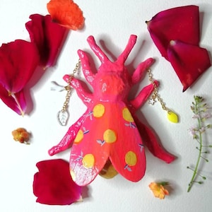 Puede incluir: Un insecto de papel maché rosa con diseños de limón amarillo y detalles de cadena dorada. El insecto está rodeado de pétalos de rosa rosa y naranja.