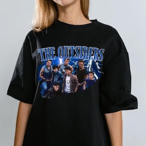 May include: Black t-shirt featuring a graphic of a group of people with the text "THE OUTSIDERS" in blue. The graphic has a vintage, retro style with a lightning bolt design in the background.