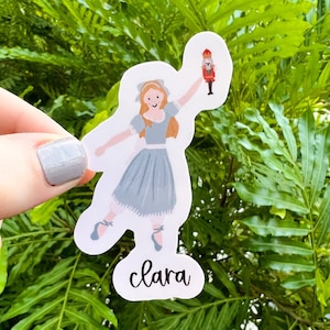 May include: A white sticker with a cartoon illustration of a girl in a blue dress holding a nutcracker. The sticker has the word "clara" written in cursive below the illustration.