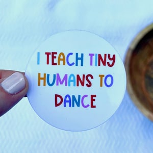 May include: White round button with the text "I teach tiny humans to dance" in colorful letters.