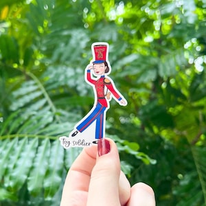 May include: A sticker of a toy soldier girl in a red and blue uniform with a red hat. The sticker is held in a hand with red nail polish. The text "toy soldier" is written below the sticker.
