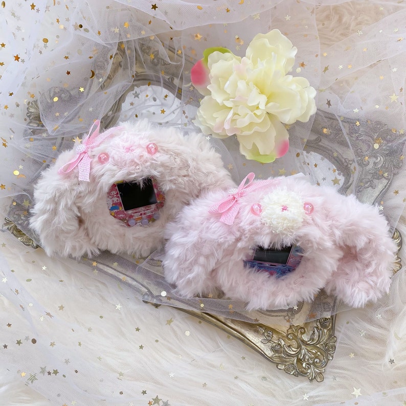 May include: Two plush, light pink and white, character-shaped cases with a screen. Each case has a pink bow and a small, decorative flower. The cases are on a decorative, gold-colored tray, with a white and gold background.