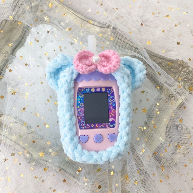 May include: A light blue, plush, crocheted case for a handheld electronic device, shaped like a bubble tea cup. It features a pink bow with a pearl accent and a pink and purple device with a screen and buttons. The device has Chinese text.