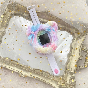 May include: A white Tamagotchi watch with a fluffy, pastel-coloured bear-shaped cover and a light blue bow. The watch has a small screen and a white strap. The background is a gold-framed mirror and white fabric with gold stars.