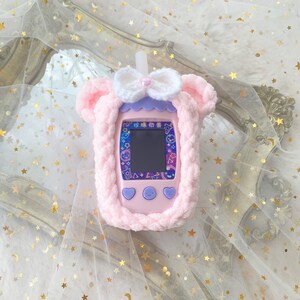 May include: A pink handheld electronic game console with a screen, buttons, and a white straw. It is encased in a soft, light pink crocheted cover with bear ears, a white bow, and a purple trim. The screen displays colorful graphics.