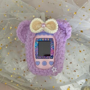 May include: A lilac-colored handheld electronic game device with a screen, buttons, and a protective cover. The cover is made of soft, fuzzy material and features a cream-colored bow with a pearl accent. The device has Japanese text on the screen.