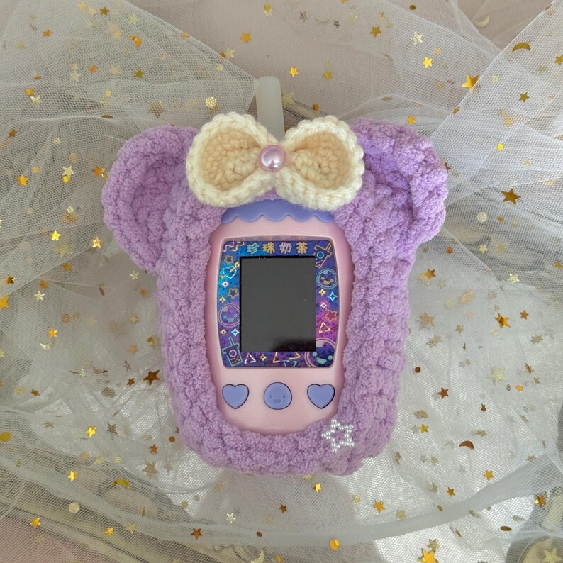 May include: A lilac-colored handheld electronic game device with a screen, buttons, and a protective cover. The cover is made of soft, fuzzy material and features a cream-colored bow with a pearl accent. The device has Japanese text on the screen.