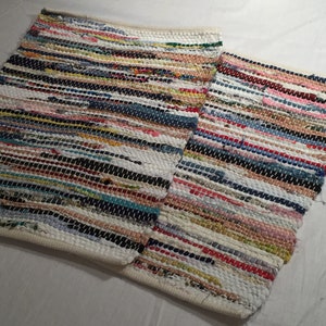 May include: Two rectangular rag rugs with a woven design. The rugs feature a mix of colorful fabric strips in various shades, including blue, red, yellow, and white. The edges are finished with a white border.